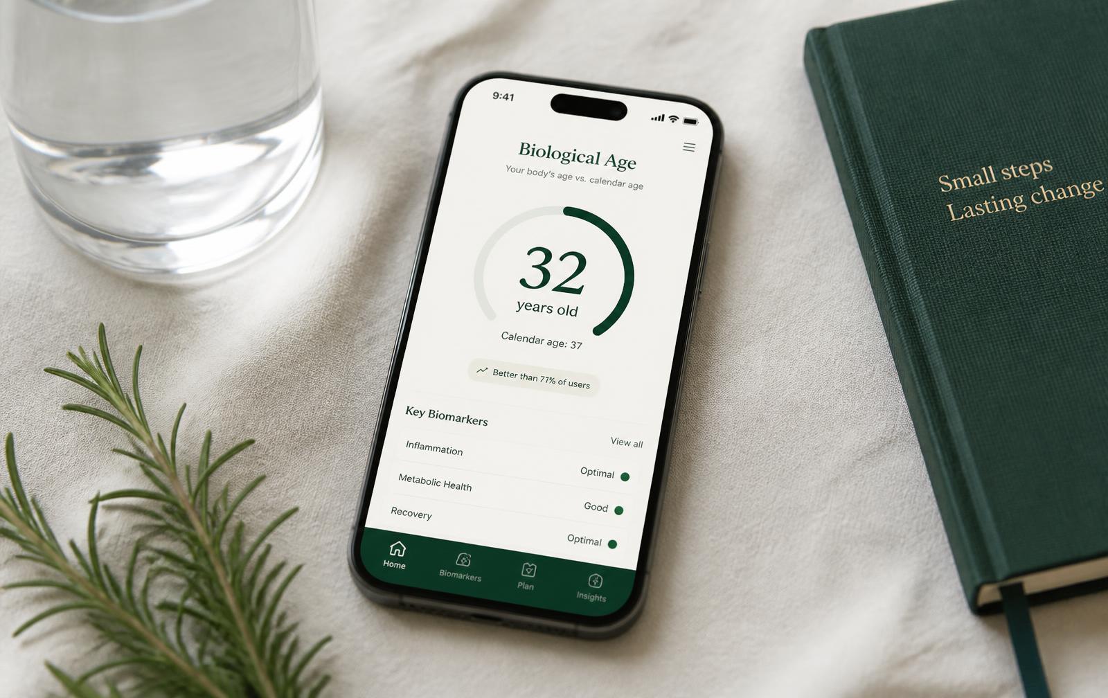 Smartphone on cream linen showing an emerald biological age dashboard with a score ring, beside a green notebook, rosemary and a glass of water